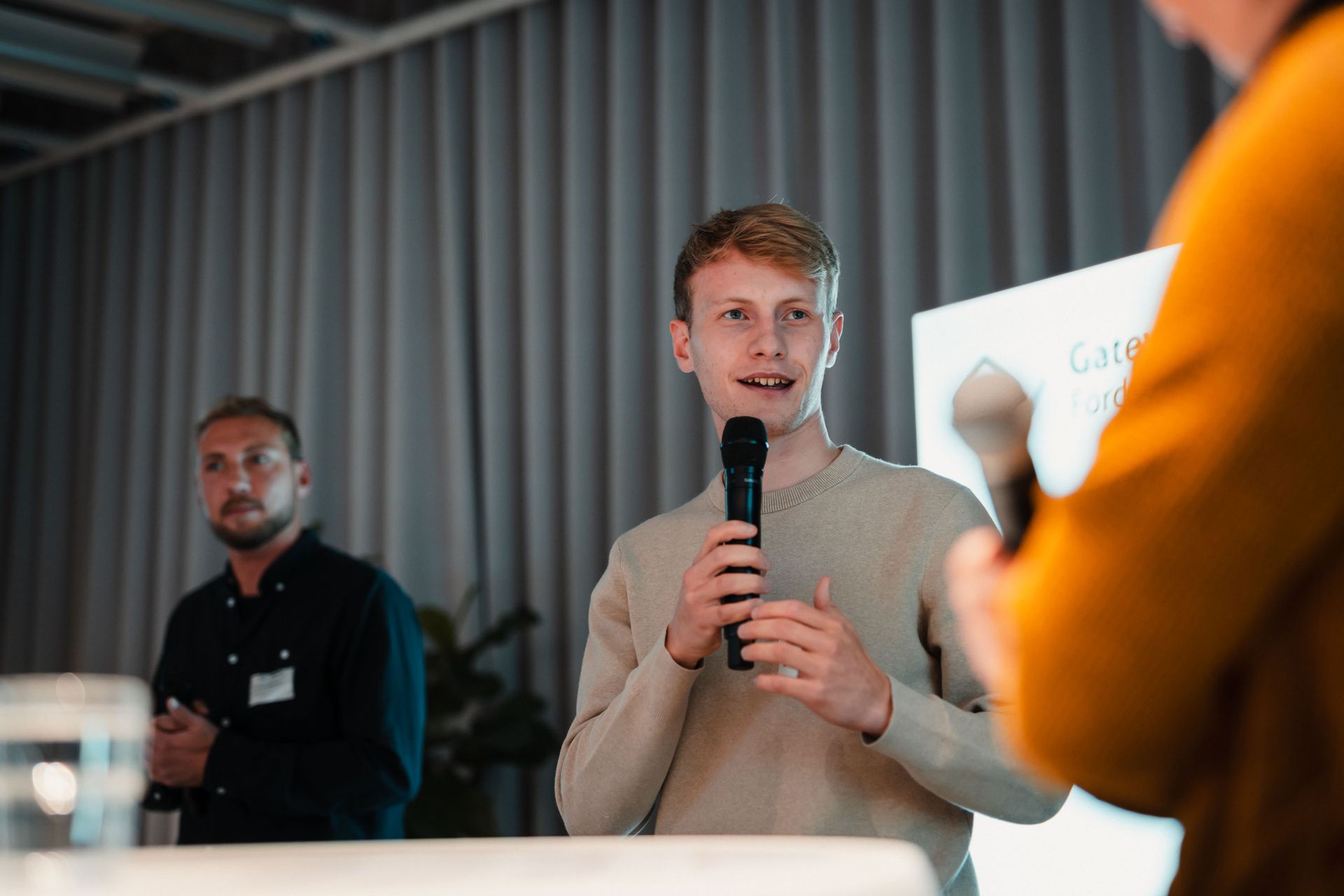 Ein Mann spricht mit einem Mikrofon bei einer Podiumsdiskussion, im Hintergrund steht ein weiterer Mann.