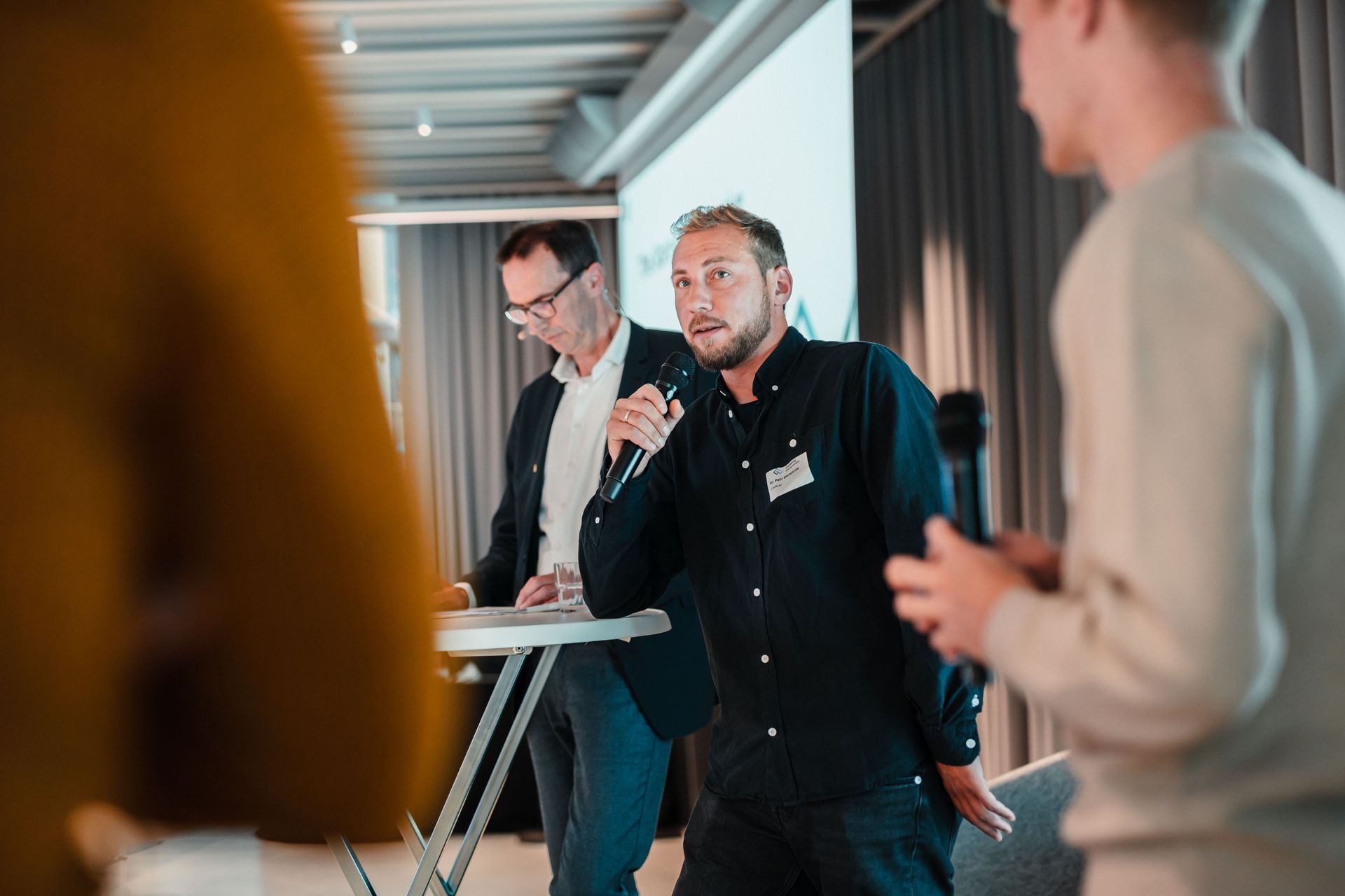 Ein Mann spricht mit einem Mikrofon bei einer Podiumsdiskussion, im Hintergrund steht ein Mann an einem Stehtisch.