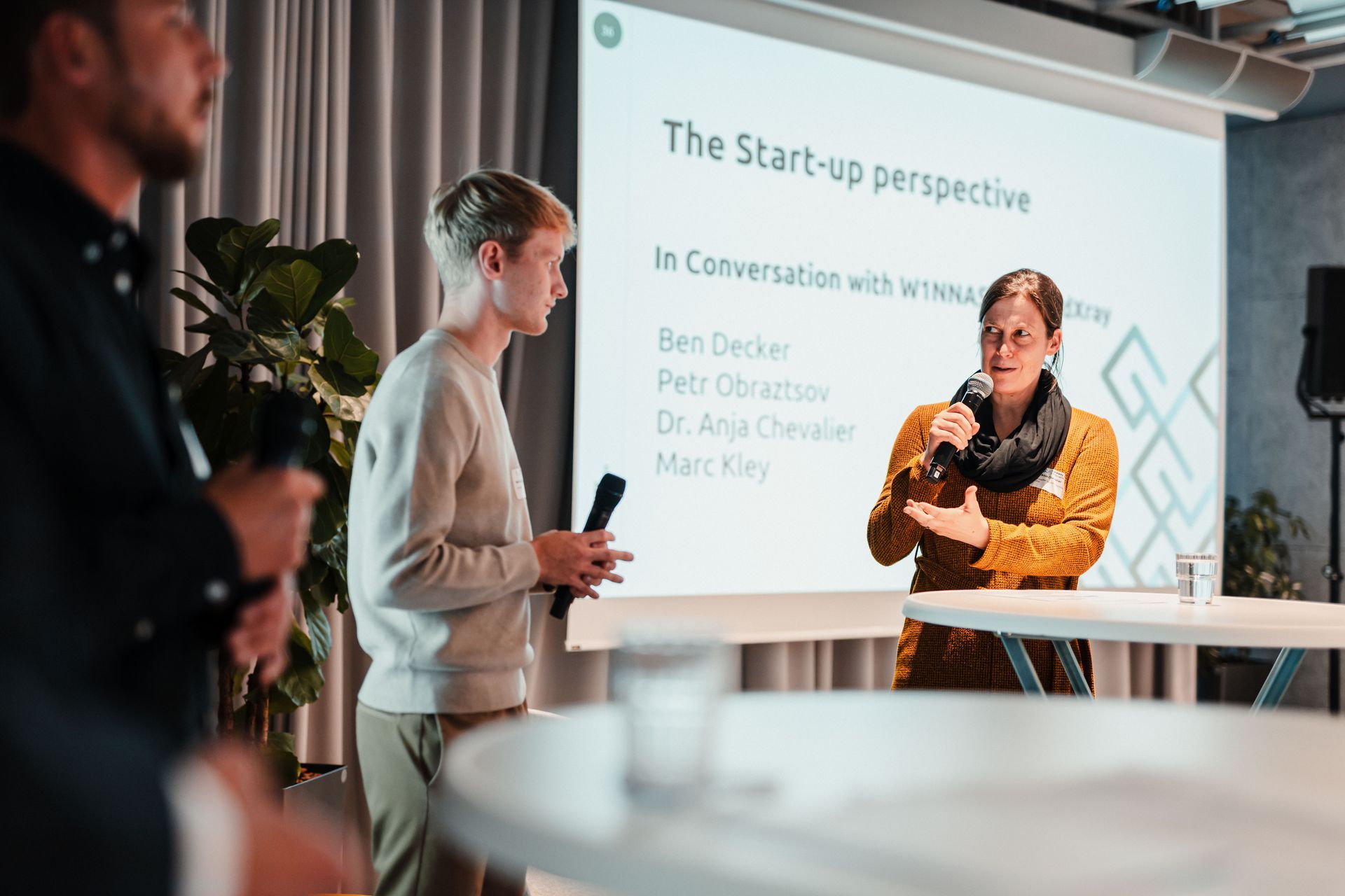 Mehrere Personen führen auf einer Bühne ein Gespräch mit Mikrofonen; im Hintergrund ist eine Präsentationsfolie mit dem Titel „The Start-up perspective“ zu sehen.