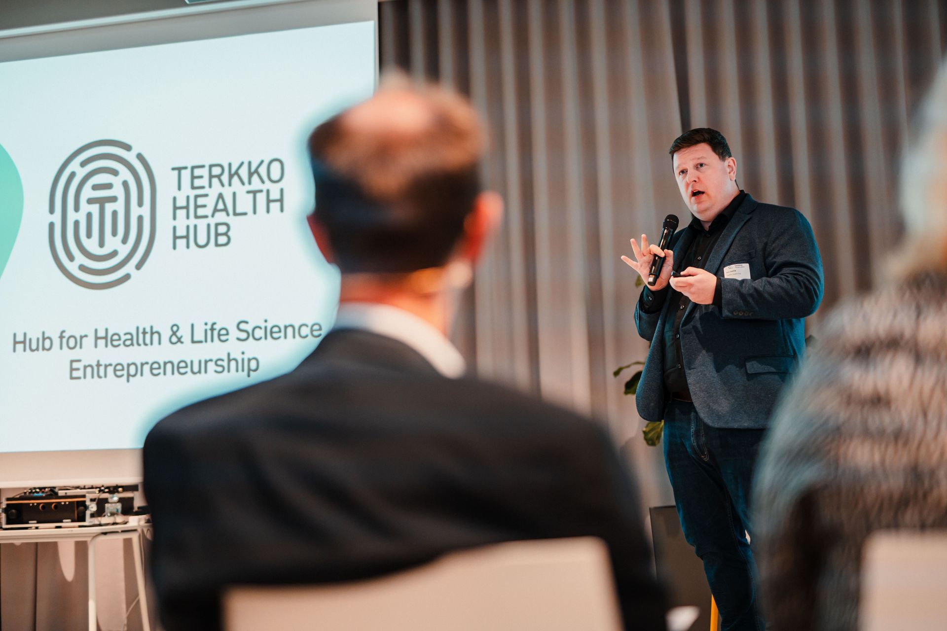 Ein Sprecher präsentiert mit Mikrofon vor einer Leinwand, auf der das Logo und der Name „Terkko Health Hub“ angezeigt werden; im Vordergrund sitzt Publikum.