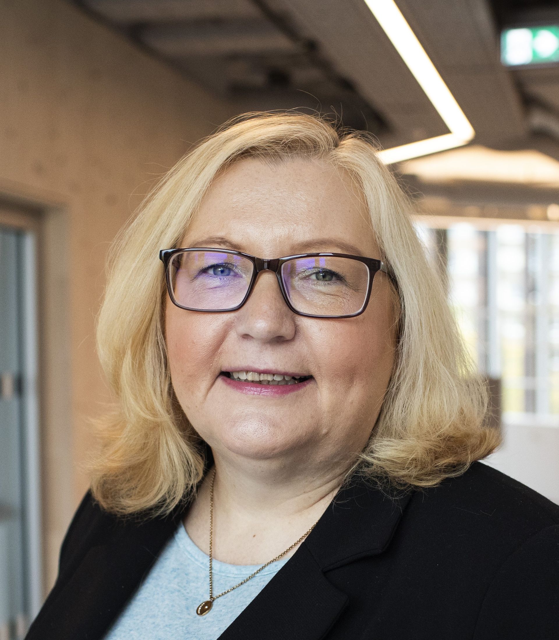 Eine Frau mit schulterlangen blonden Haaren und Brille, die einen schwarzen Blazer und darunter ein weißes Oberteil trägt. Sie steht in einem hellen Raum mit großen Fenstern im Hintergrund und lächelt freundlich.