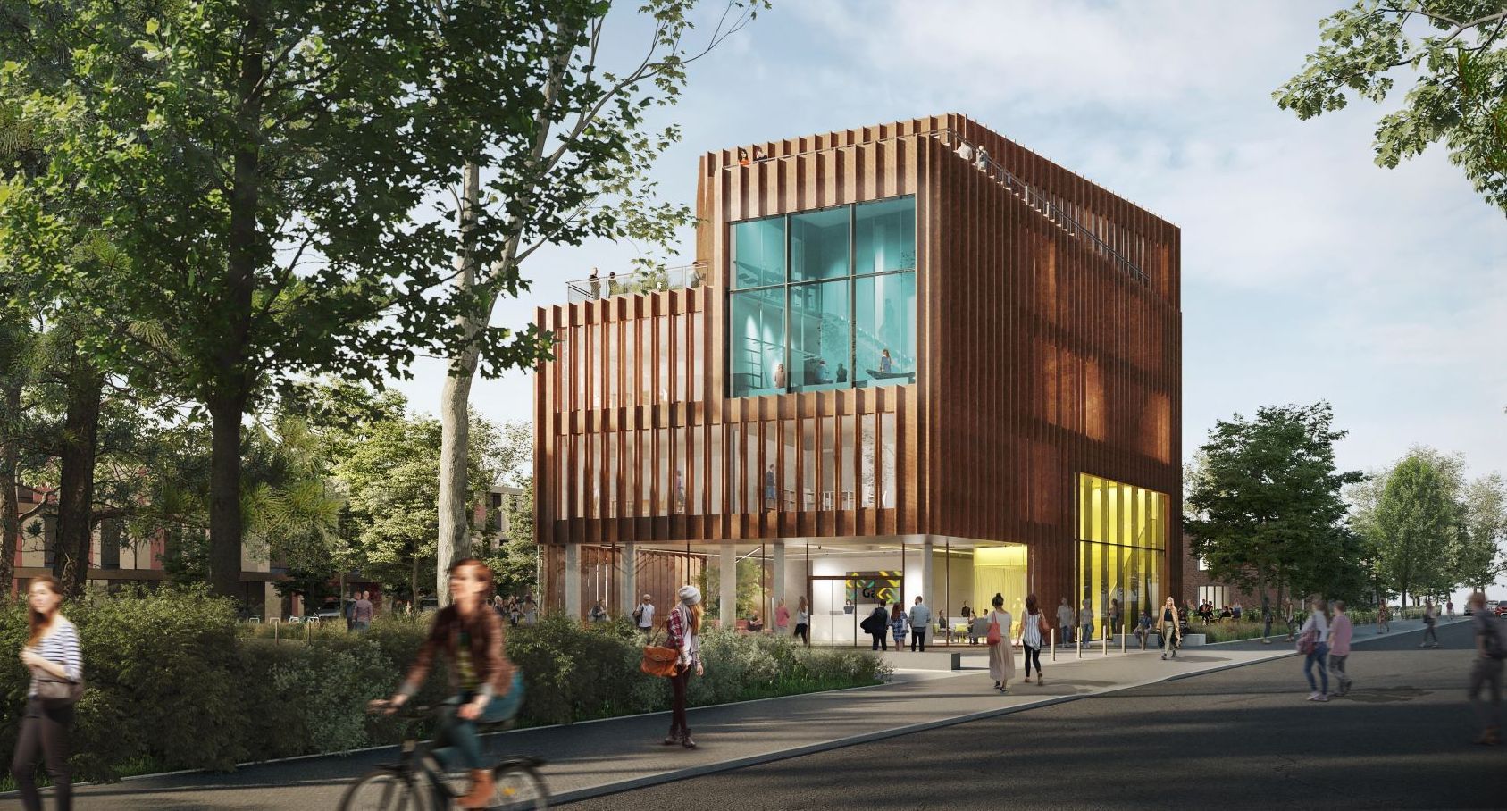 Ein modernes, würfelförmiges Gebäude mit einer Fassade aus vertikalen, rostbraunen Metalllamellen steht an einer von Bäumen gesäumten Straße. Große Glasflächen geben den Blick ins helle Innere frei. Das Gebäude hat mehrere Etagen, eine Dachterrasse und wirkt freundlich und offen. Vor dem Gebäude fahren mehrere Personen mit Fahrrädern, andere gehen zu Fuß. Im Vordergrund ist ein grüner Streifen mit Büschen und Bäumen zu sehen, der eine angenehme, städtisch-natürliche Atmosphäre schafft.