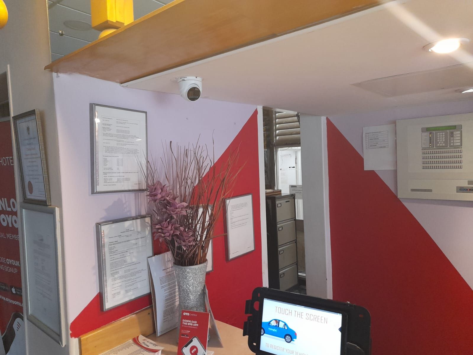 Hikvision camera mounted in Chiltern hotel reception area with framed certificates and a red accent wall.