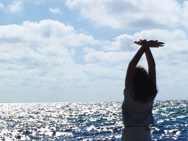 Luz, mar, niña de espaldas alzando los brazos al cielo.
