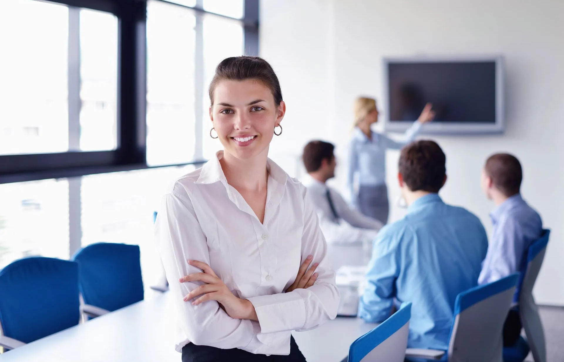 Una donna in primo piano e delle persone sedute intorno a una scrivania che guardano uno schermo, in un ufficio.