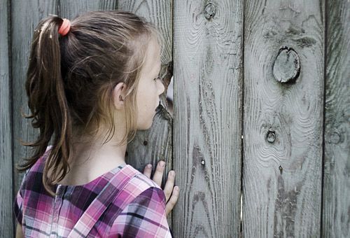 Durch Neugier näher ans Ziel kommen. Mädchen späht durch ein Astloch in einem Bretterzaun. Neugierde, Entdeckerfreude.