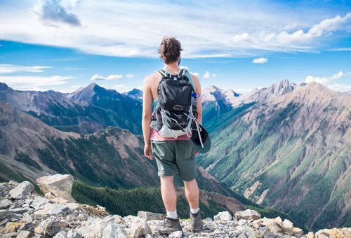 Angstbewältigung. Link zu Terminbuchung. Mann steht auf einem steinigen Berggipfel, blickt in die grüne Ebene und auf das Bergpanorama, Freiheit.