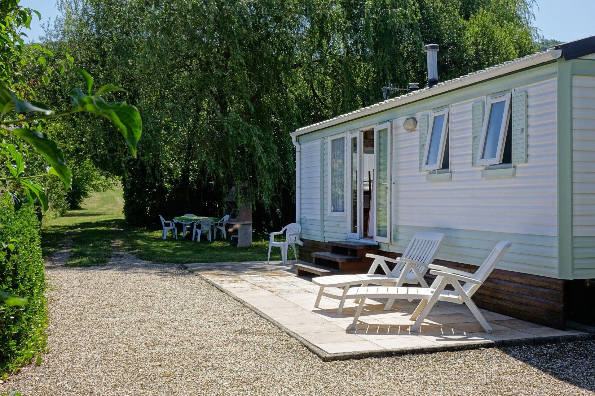 Mobile-home, vue de la terrasse avec deux transats blancs, chemin en graviers et table de jardin avec chaises