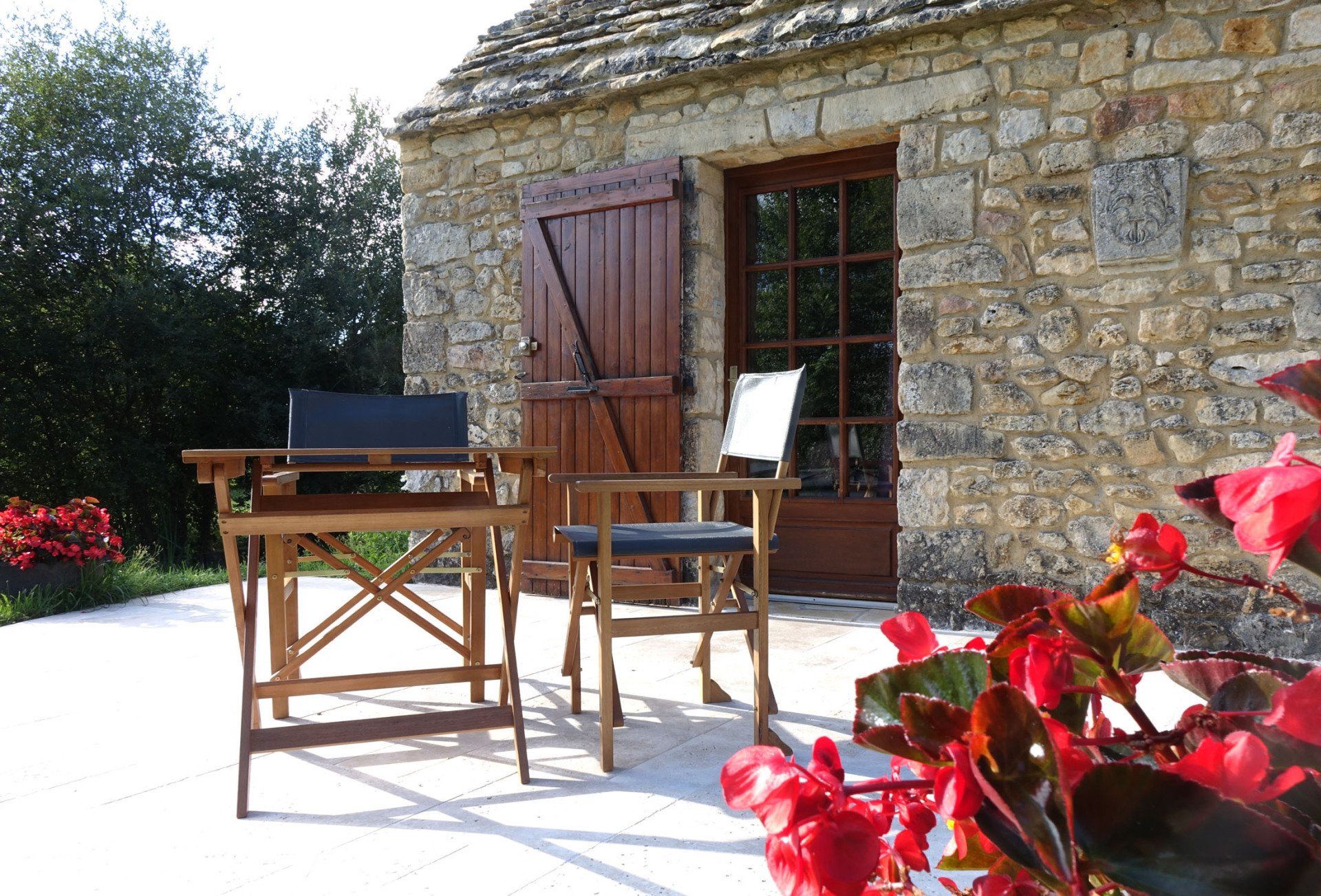 Vue sur la chambre La Borie (petite maison en pierres). Au premier plan table et chaises en bois et fleurs rouges