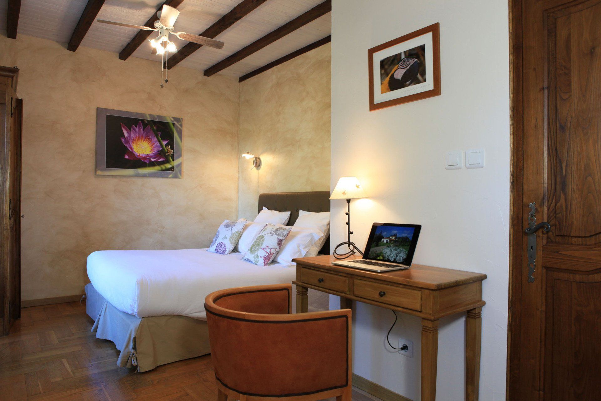 Vue intérieure de la chambre Sérénité. Fauteuil et petit bureau avec lampe de chevet et ordinateur portable - Lit avec couette blanche, photos murales