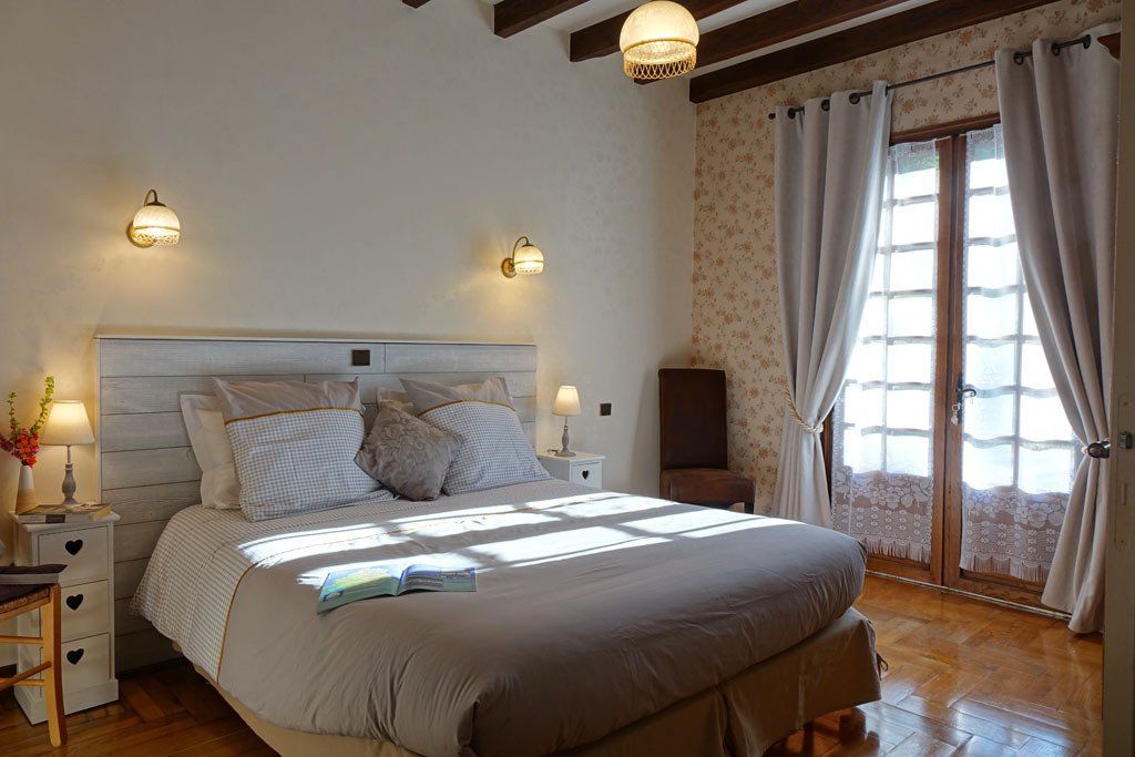 Vue intérieure de la chambre Romance, Lit et sa couette imprimée, chevets et lampes de chevets, fauteuil, porte fenêtre donnant sur l'extérieur