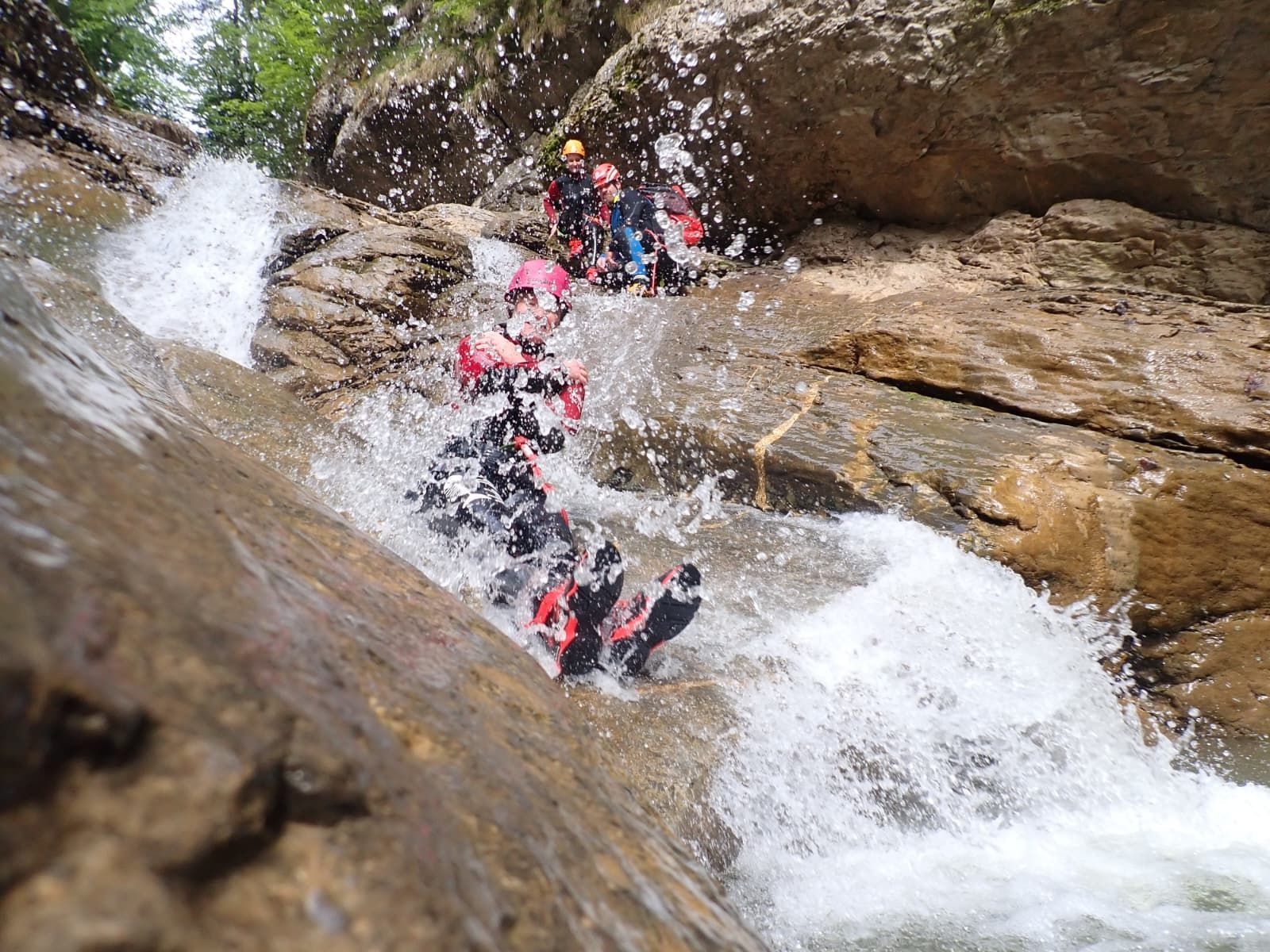 Teilnehmer der Canyoningtour in der Starzlachklamm rutschen über eine natürliche Felsrutsche durch spritzendes Wildwasser in einer engen Schlucht.