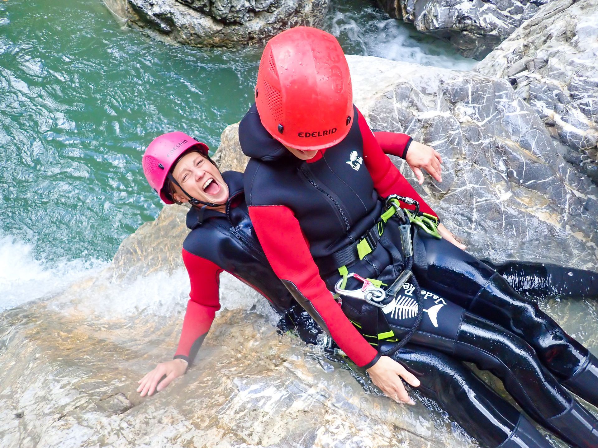 Zwei Teilnehmer der Merlins-World Canyoningtour rutschen gemeinsam über eine Felsrinne ins türkisfarbene Wasser, gesichert und ausgerüstet mit Neoprenanzügen, Helmen und Klettergurten.