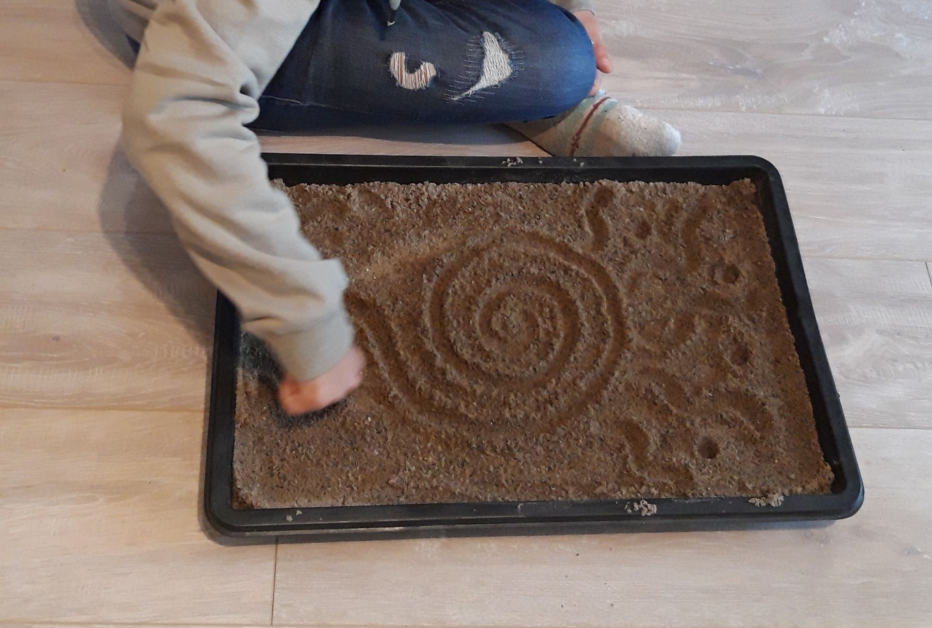 Eine Person, die mit dem Finger eine Figur in ein Blech mit Sand malt