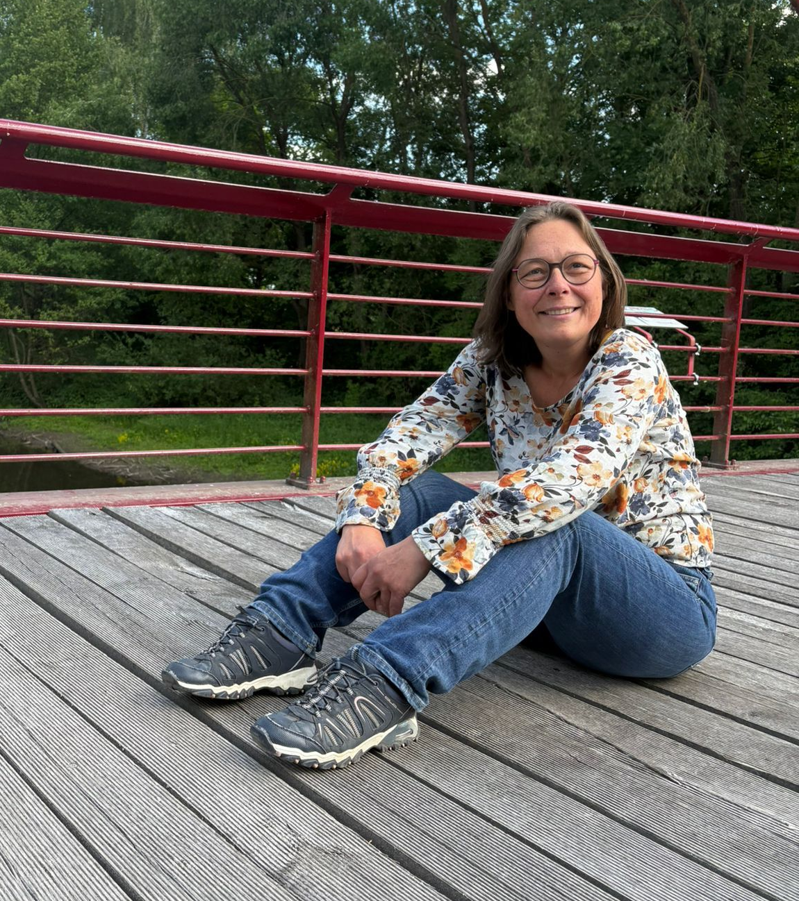 Eine Frau auf einer Holzbrücke sitzend und verträumt lächelnd in den Himmel blickt