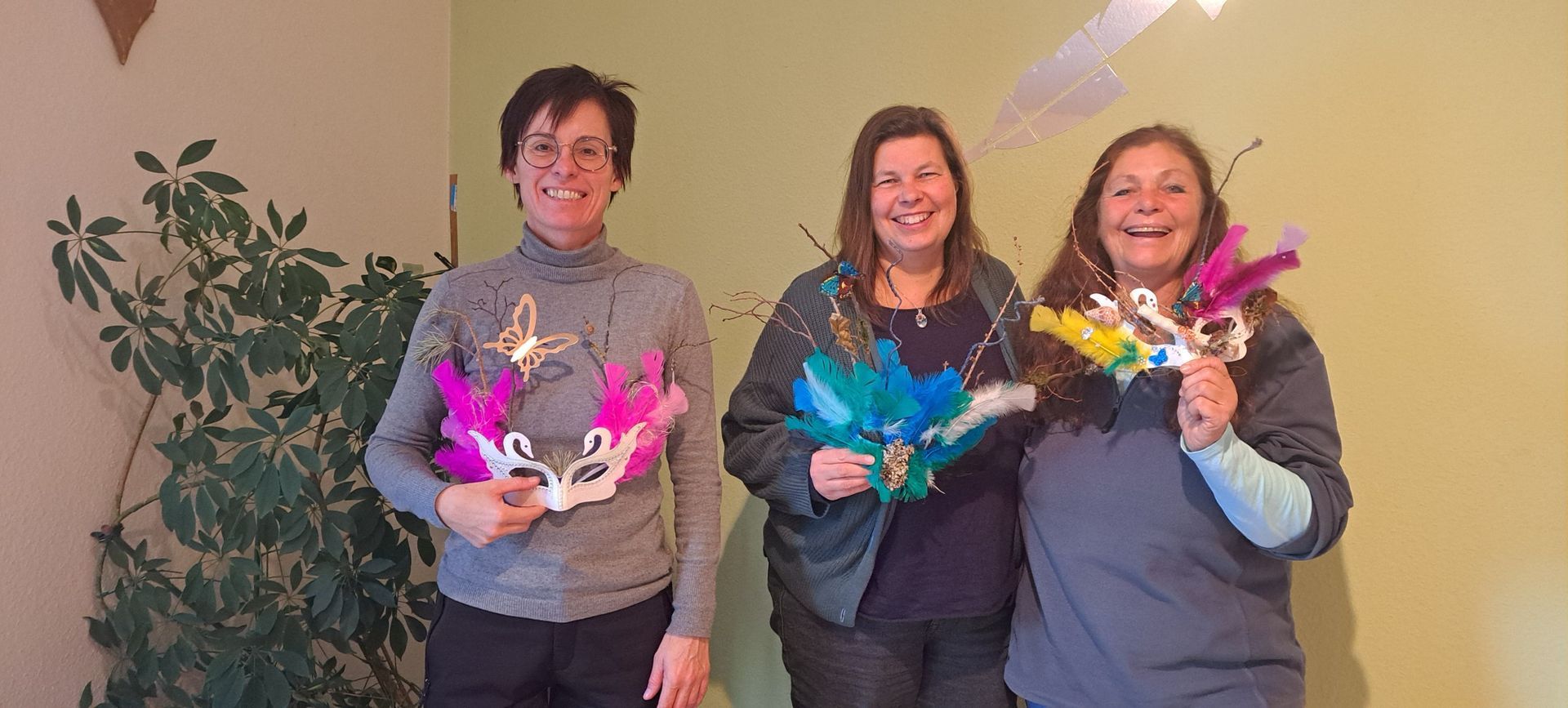 Drei lächelnde Frauen mit selbstgebastelten Masken, die sie vor sich halten
