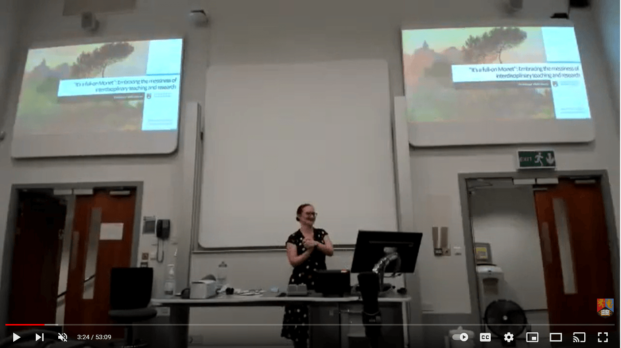 A white woman in a black dress standing at the front of a lecture theatre with two screens behind her
