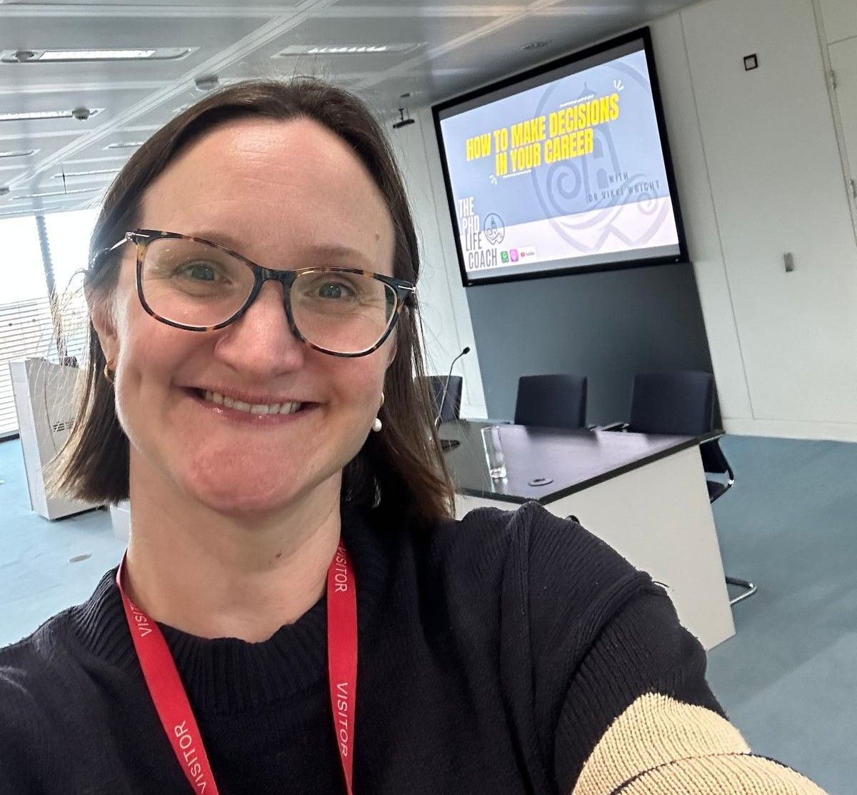 Picture of Vikki (white woman, brown bobbed hair and glasses) grinning in a seminar room with a big blue powerpoint behind her