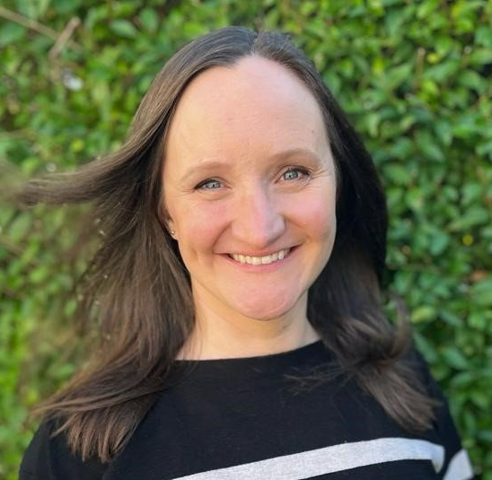 Dr Vikki Burns Woman with long dark hair smiling at the camera in front of a green hedge