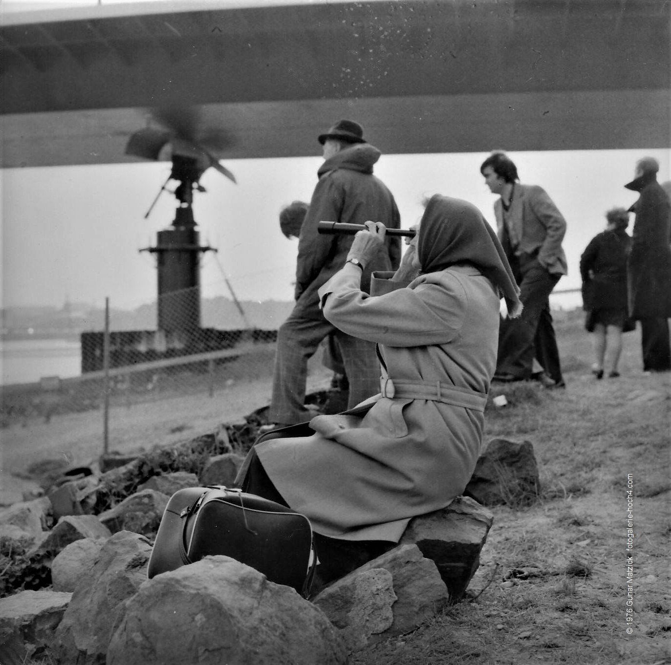 Das Ereignis aus sicherer Entfernung beobachten. Brückenverschiebung 1976. Spektakuläre Verschiebung der Oberkasseler Brücke in Düsseldorf.
Am 7. und 8. April wurde der komplette Oberbau der Brücke samt Pylon und Schrägseilen mit einem Gesamtgewicht von 12.500 Tonnen um 47,5 Meter stromabwärts verschoben (!). Um dem Trubel ein wenig zu entgehen, hatte ich das Ereignis linksrheinisch auf den Oberkasseler Wiesen beobachtet.
Dieser Schnappschuss war mir aus meiner Sicht echt gelungen.
Die Dame im Trenchcoat, ihre Brille akkurat auf die Tasche gelegt und mit ihrem Teleskop das Treiben entspannt beobachtend – herrlich dieser Kontrast.
Apropos Eigenlob, dieses Foto brachte mir bei dem gleichzeitigen Fotowettbewerb der Düsseldorfer Tageszeitung EXPRESS den 1. Platz. Gewonnen hatte ich übrigens meine erste Nikon und ein Fässchen Gatzweilers Alt.
Wie komme ich eigentlich jetzt darauf? Ich war 2017 im NRW-Forum Düsseldorf auf der Fotoausstellung von Nikon: I AM 100! Ja, Erinnerungen wurden wach. Düsseldorf-Oberkassel, NRW, Deutschland.