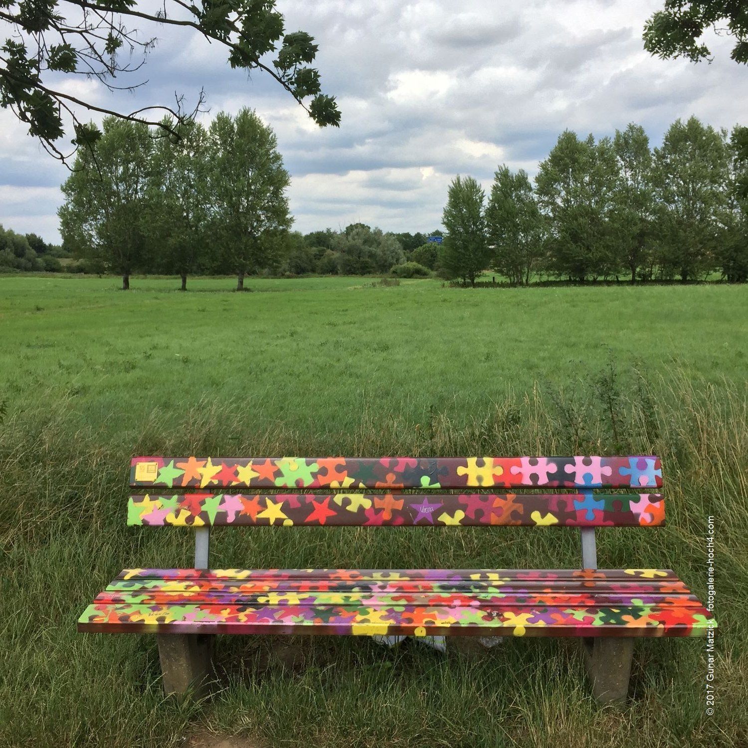 Bunt wie die Welt. Bunt wie die Welt. Herzlich willkommen und nehmt doch Platz. Wer kann dieser fröhlichen Einladung widerstehen? Sterne, Puzzleteile, Regenbogenfarben - eine so herrliche positiv (un-) besetzte Bank im Grünen. Lass dich tragen, einstimmen, animieren. Einfach mitmachen. Leverkusen, NRW, Deutschland.