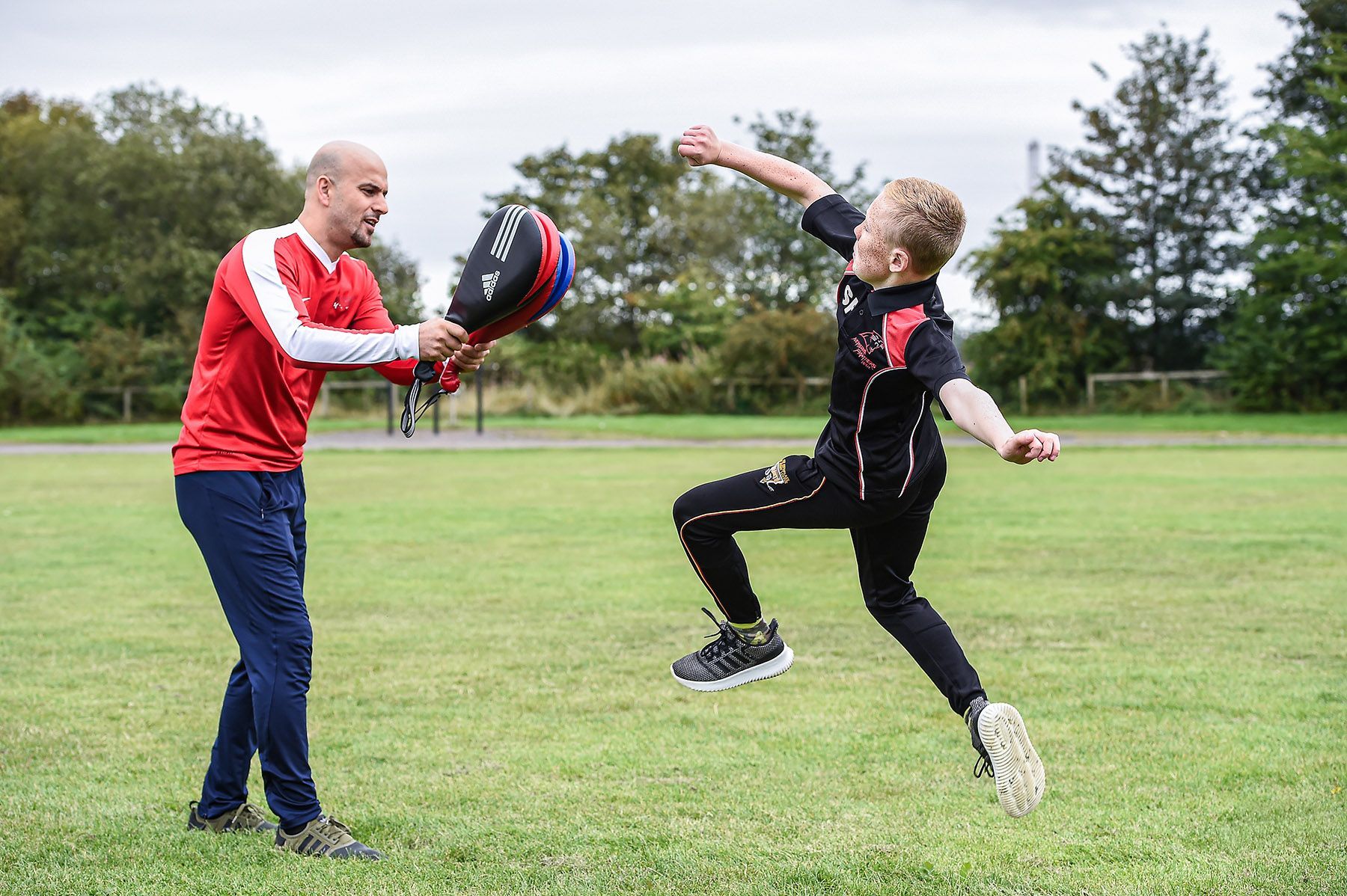 A young boy is doing a martial arts kick onto pads that his trainer is holding.