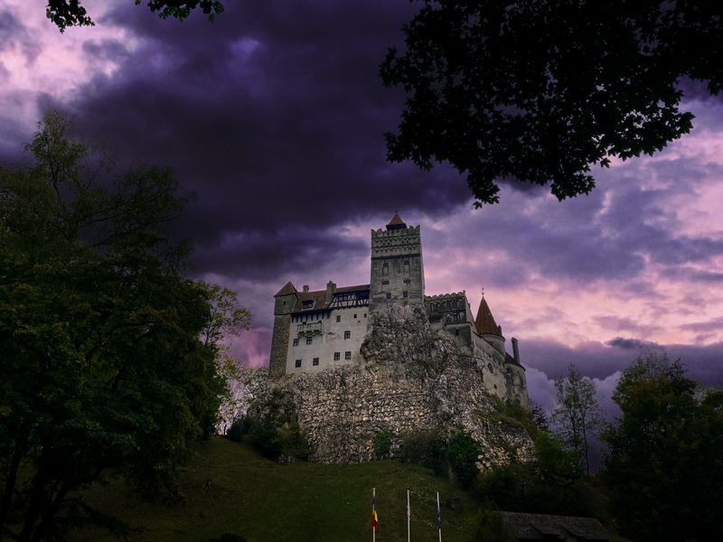 Das Schloss Bran in Rumänien erhebt sich auf einem Felsen, umgeben von dunklen Wäldern und dramatischem, wolkenverhangenem Himmel.