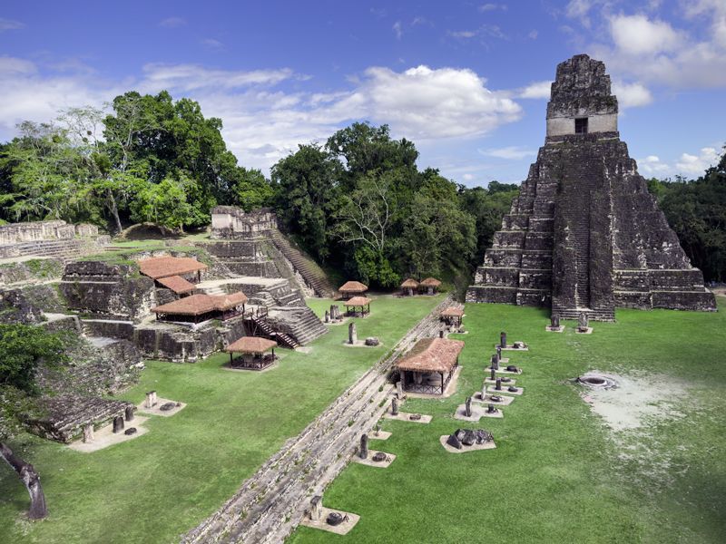 Der große Maya-Tempel I von Tikal ragt zwischen dichtem Regenwald in den Himmel; im Vordergrund liegen antike Ruinen und Lichtungen.