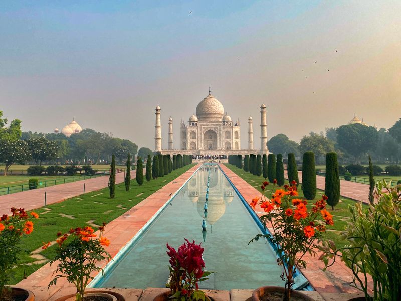 Frontalblick auf das Taj Mahal bei weichem Morgenlicht, mit reflektierendem Wasserbecken und symmetrischen Gartenwegen im Vordergrund.