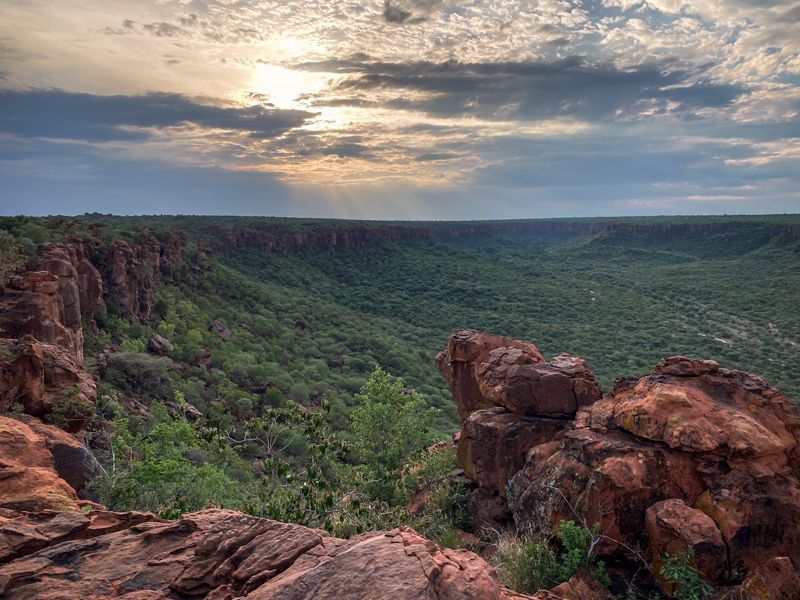 Sonnenuntergang über rötlichen Felsformationen in Namibia; im Vordergrund ein ausgedehnter Canyon mit trockenem Buschland.