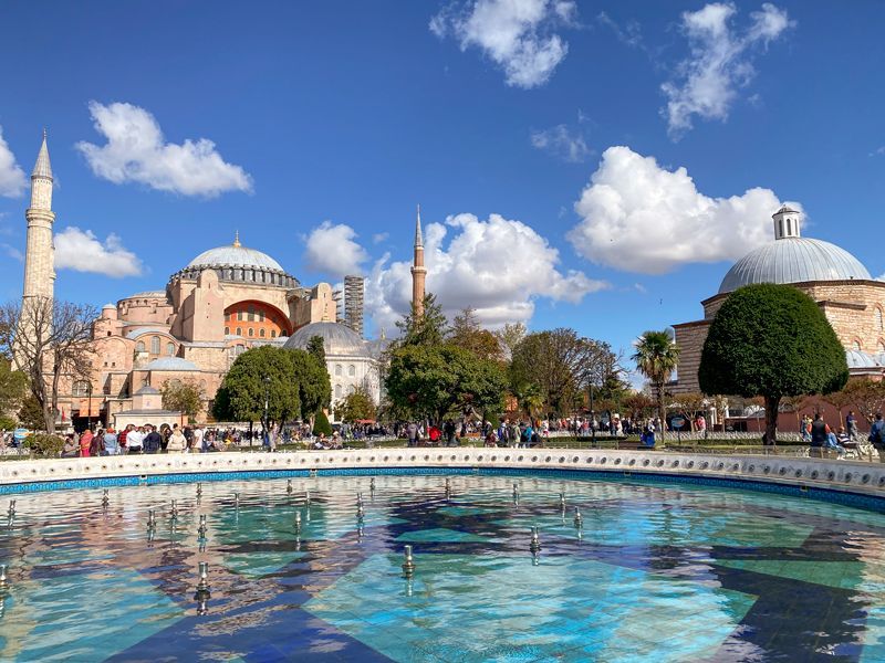 Die Hagia Sophia in Istanbul bei blauem Himmel, mit einem davorliegenden Springbrunnen und Spaziergängern im Park.