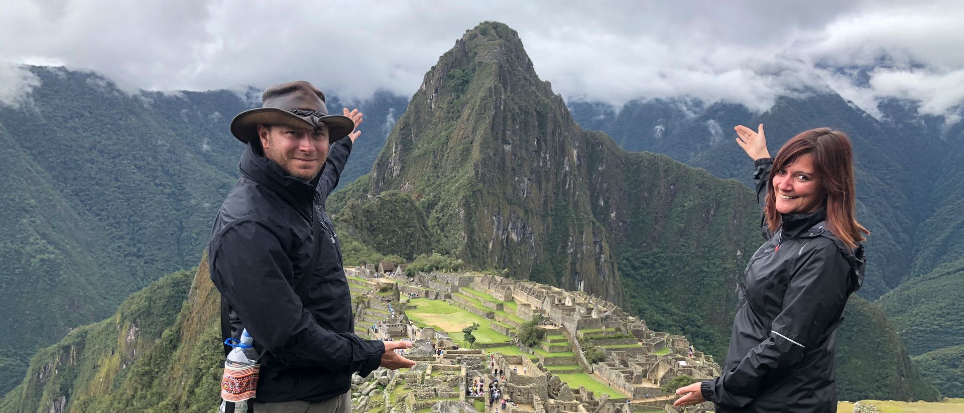 Matthias Brugger und Sandra Brugger vor Machu Picchu Ein Paar steht auf einer Aussichtsplattform vor Machu Picchu und zeigt mit den Händen auf die berühmte Inka-Stätte und die umgebenden Berge.