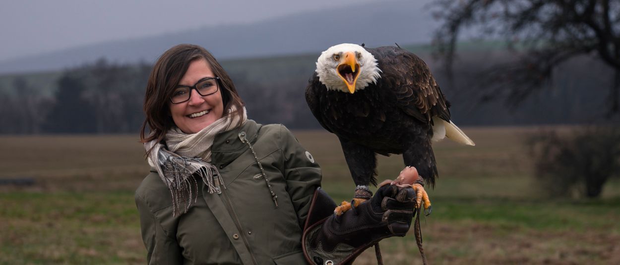 Sandra Brugger & Weißkopfseeadler Sandra Brugger im Outdoor-Outfit hält einen Weißkopfseeadler auf einem Falknerhandschuh; der Adler hat den Schnabel geöffnet.