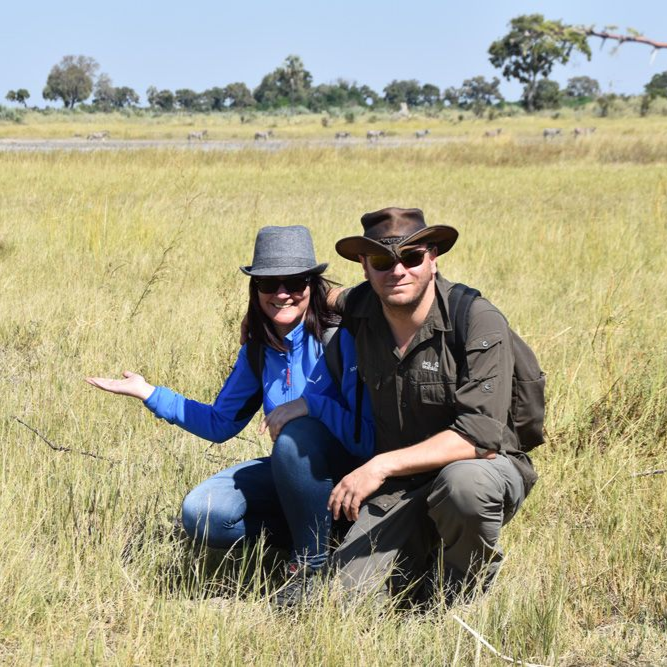 Ein Paar kniet im hohen Gras einer afrikanischen Landschaft; die Frau deutet lächelnd auf die Umgebung