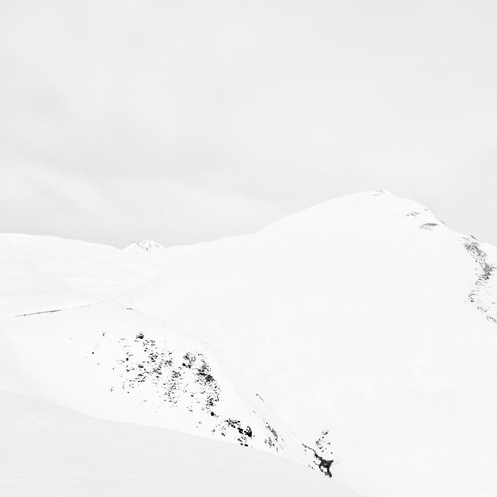 There is human beings - Il y a toujours des êtres humains : série de photographies noir et blanc épurées de paysage de montagne sous une neige blanche et pure, avec quelques traces de végétation ou de terre