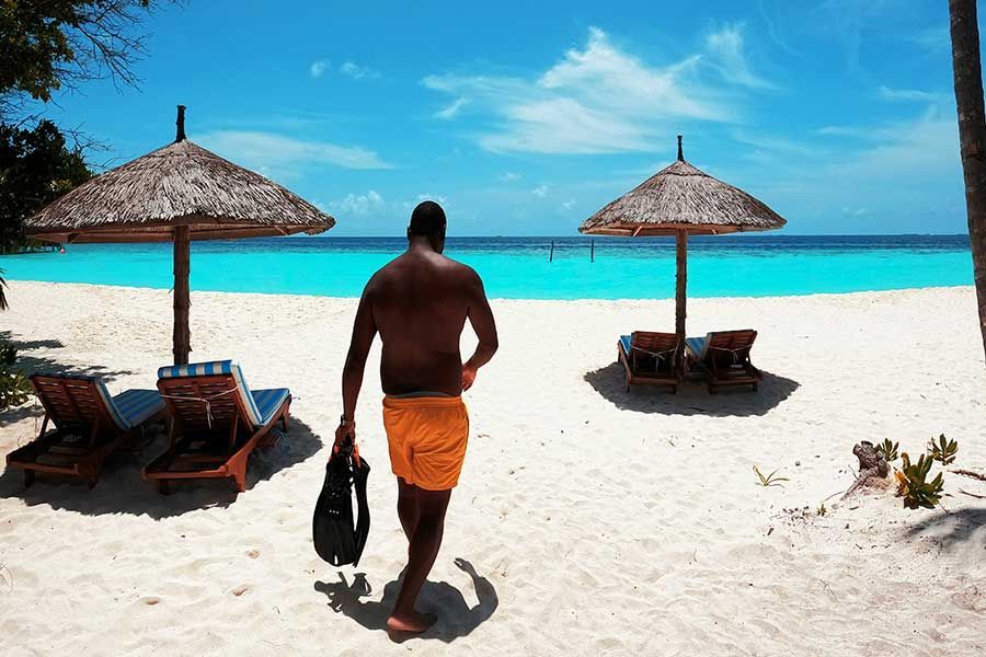 Man walking toward the water with snorkel fins, thatched umbrellas on a Maldives beach Man walking toward the water with snorkel fins, thatched umbrellas on a Maldives beach
