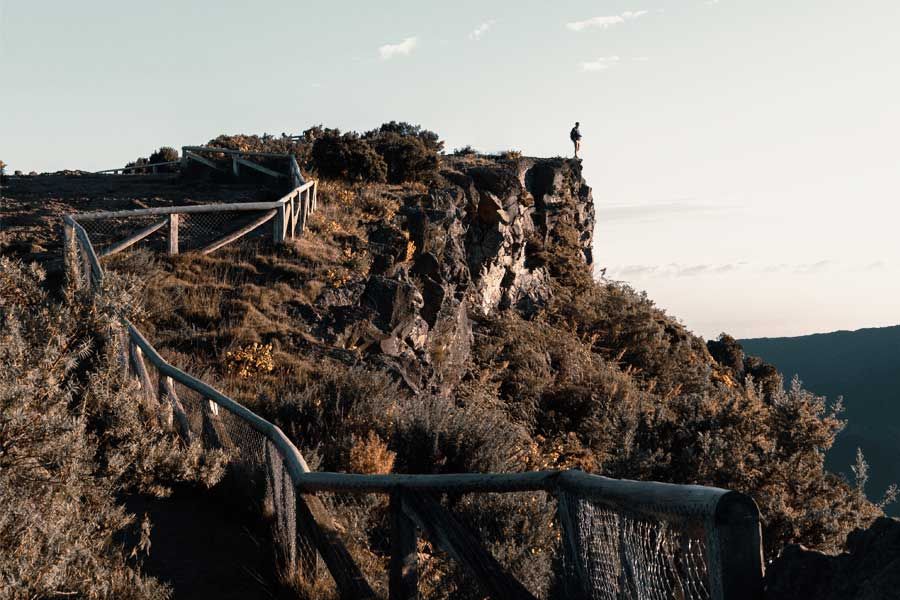 Person standing on a rocky cliff edge along a wooden fence hiking trail