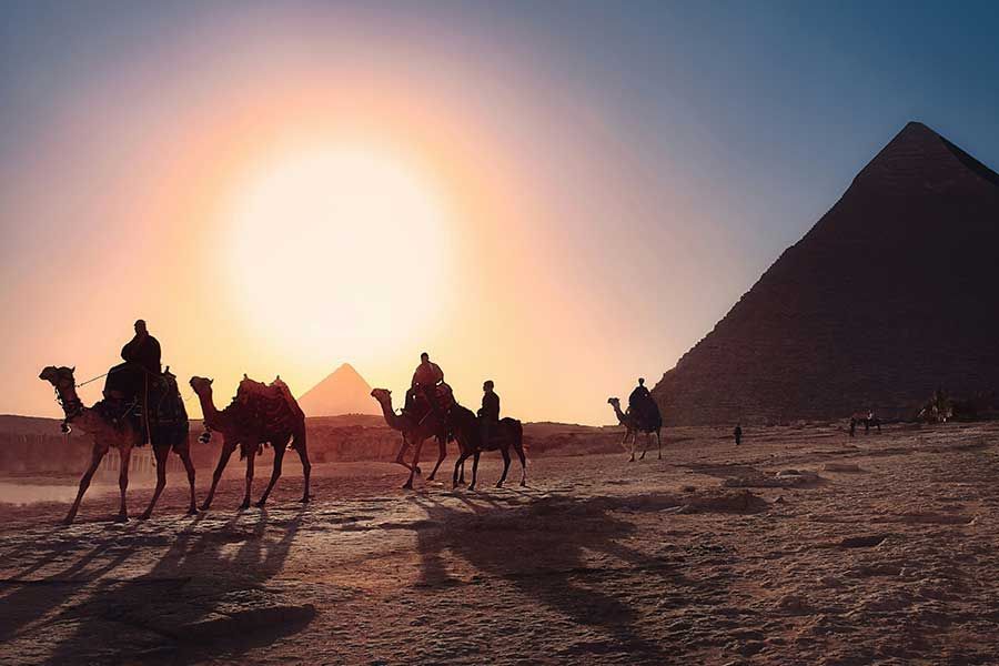 Silhouettes of camels and riders walking toward pyramids at sunset