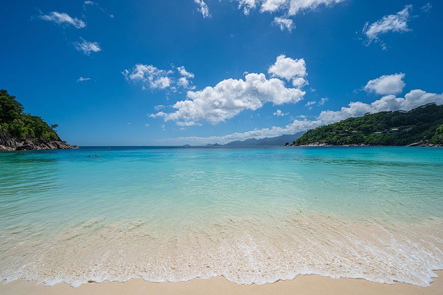Calm, shallow turquoise bay and white sand beach in the Seychelles, with green hills in the background Calm, shallow turquoise bay and white sand beach in the Seychelles, with green hills in the background
