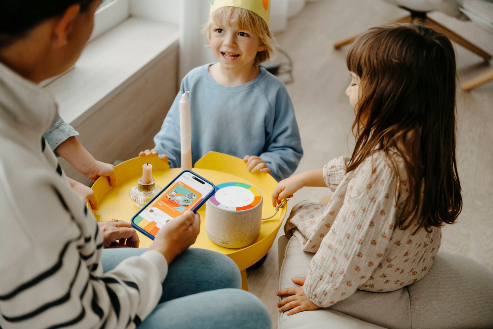 Eine Mutter bespielt mit der wobie fav-App die wobie box gemeinsam mit zwei Kindern. Einem blonden Jungen, der eine gelbe Papierkrone trägt, und einem braunhaarigen Mädchen.