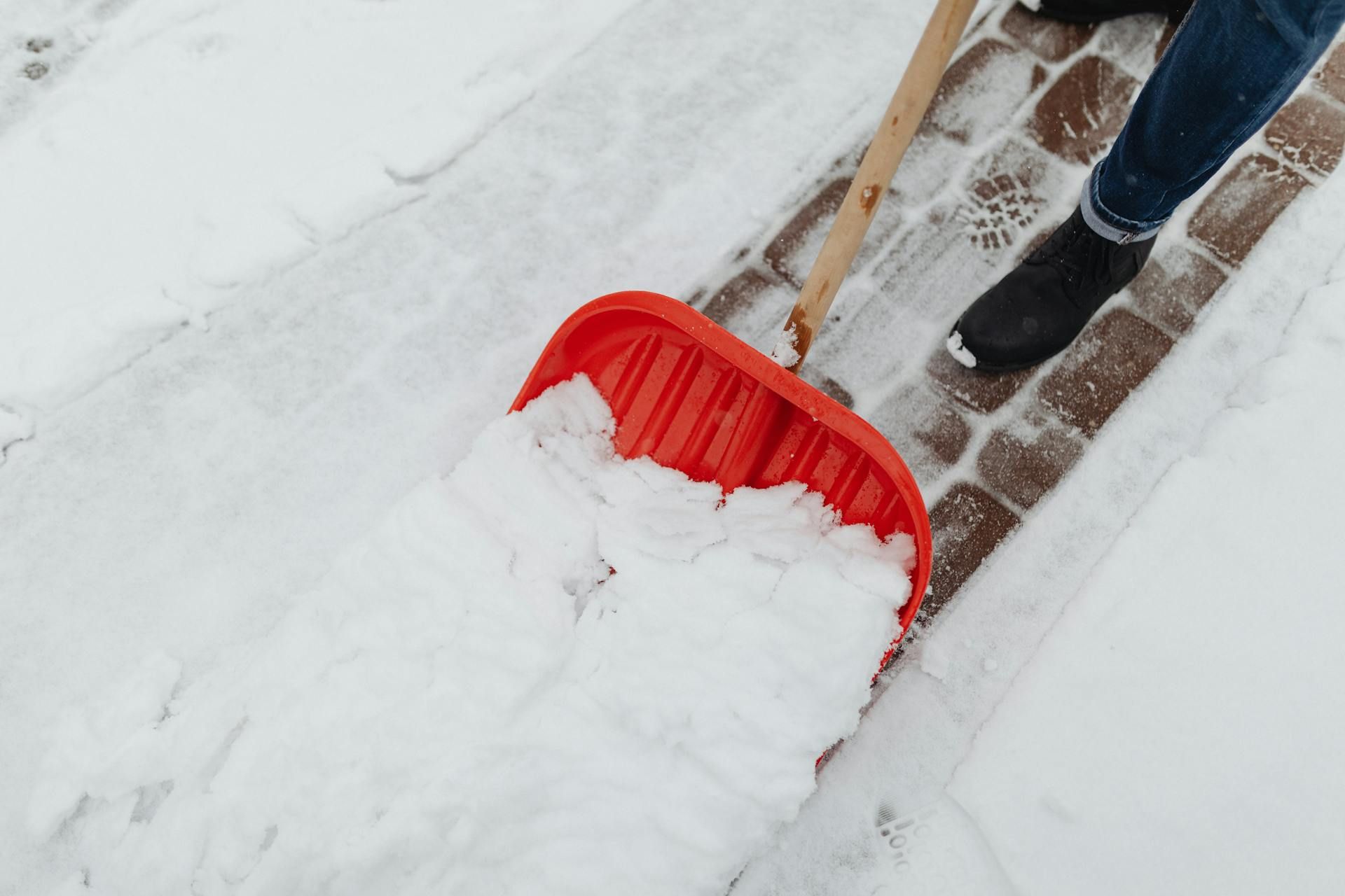 Eine rote Schneeschaufel, die auf einem gepflasterten Gehweg eine Schneise in den Schnee schiebt