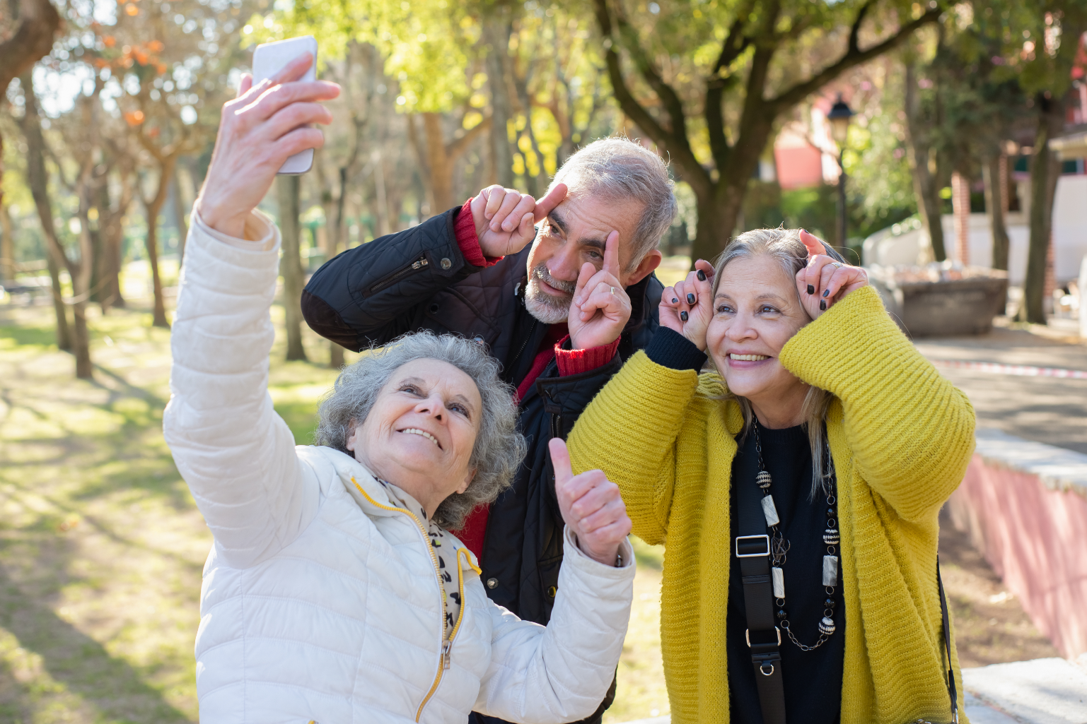 Drei ältere Menschen machen lachend ein Selfie im Park.