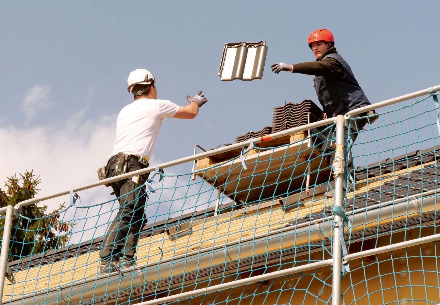 Zwei Dachdecker werfen sich auf einem Dach eine Dachpfanne zu