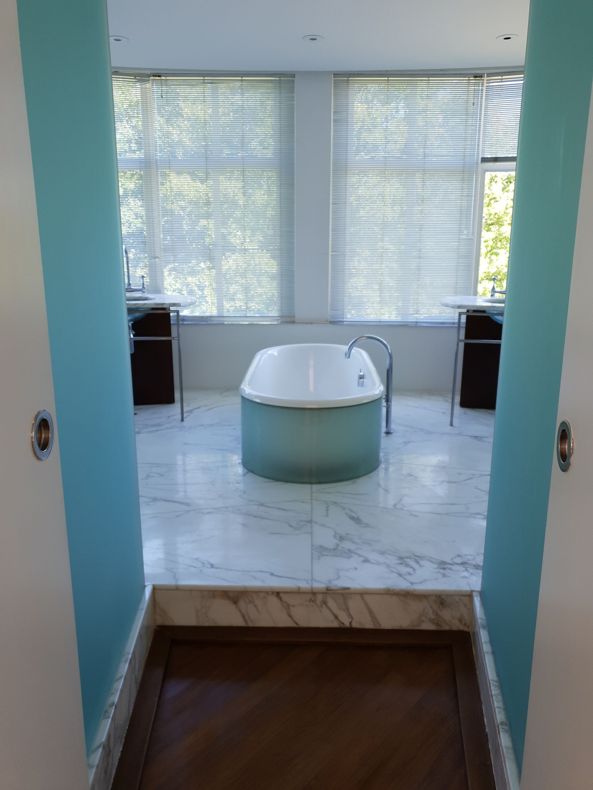 A view of a modern bathroom featuring a freestanding bathtub, surrounded by large windows with sheer blinds and light-colored walls. The floor has a mix of marble tiles and wooden accents, creating a serene atmosphere. Enhancing the room's appeal, the bathtub adds to the ambiance suitable for deep cleaning tasks, reflecting a thorough deep clean by Love Filthy Cleaning.