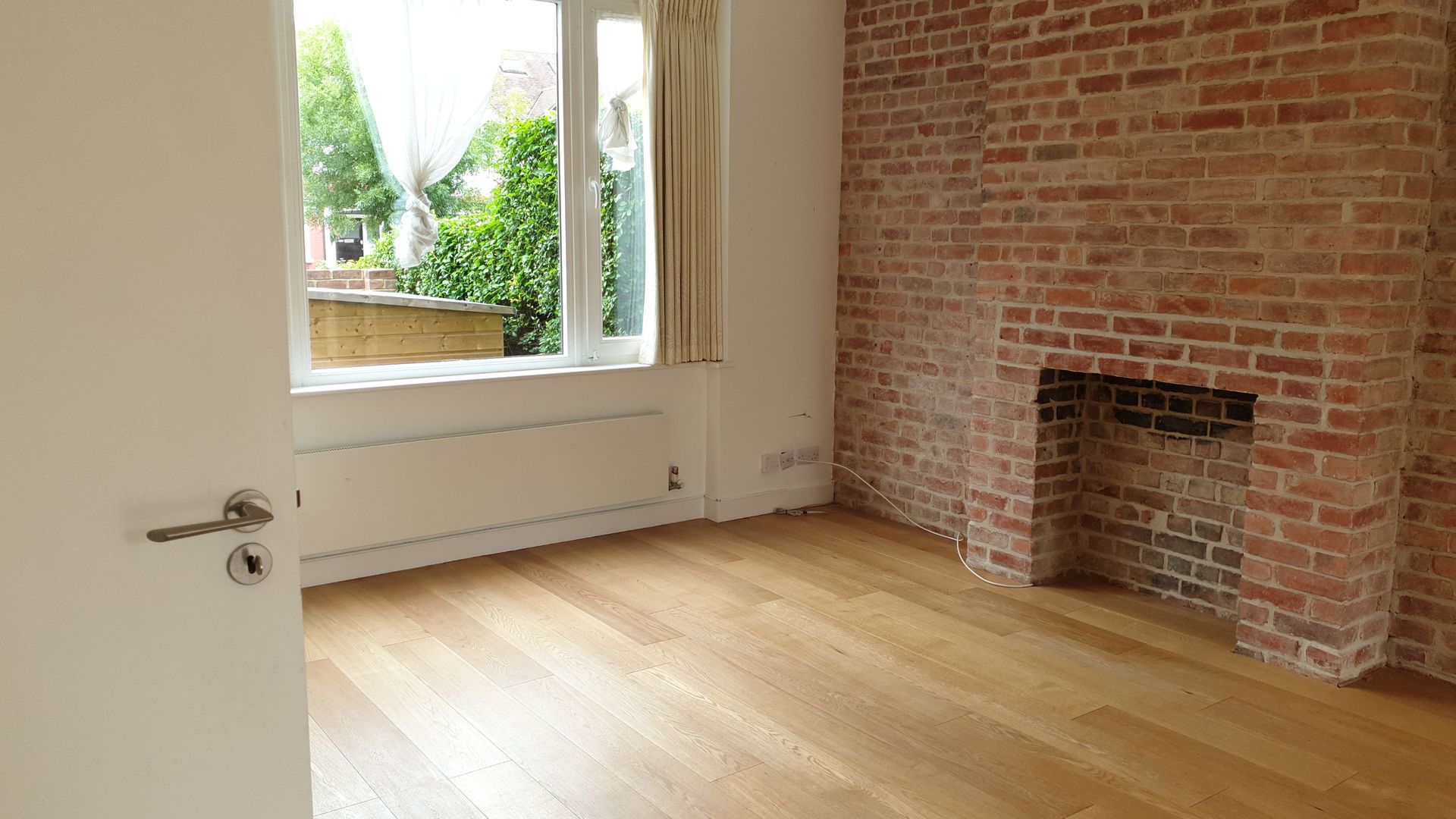 A freshly cleaned empty room after an end of tenancy clean, featuring light wooden flooring, a large window revealing greenery outside, and a brick wall with a fireplace. The slightly open white door leads into the spotless space, ready for its next occupant.