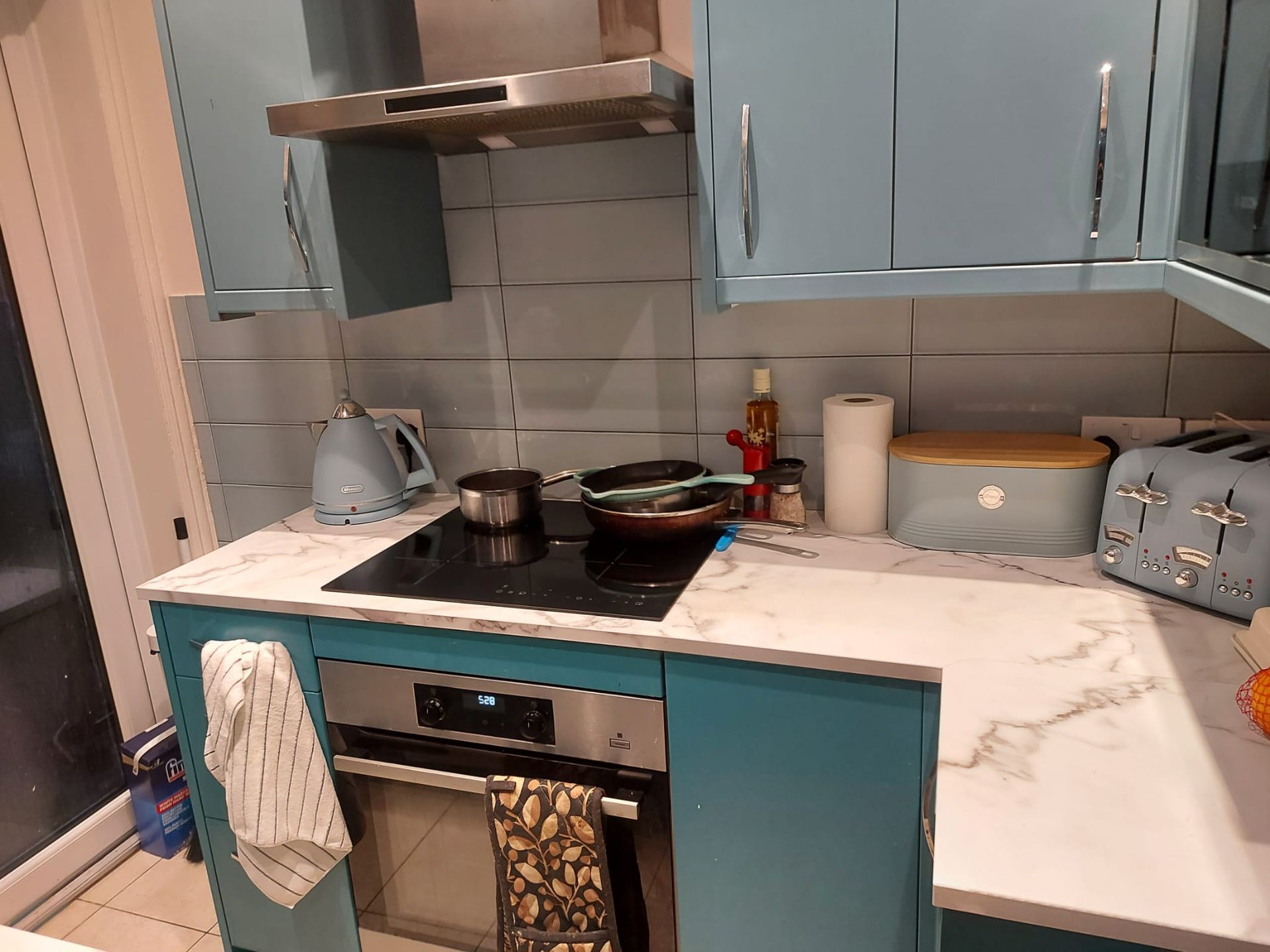 A sleek, modern kitchen with blue cabinets, a marble countertop, and an induction stove. A kettle and frying pan sit on the cooktop, while a roll of paper towels, a toaster, and a storage container are neatly arranged on the counter, enhancing the clean and organized aesthetic. The interactive slider allows users to compare the extreme cleaning results.