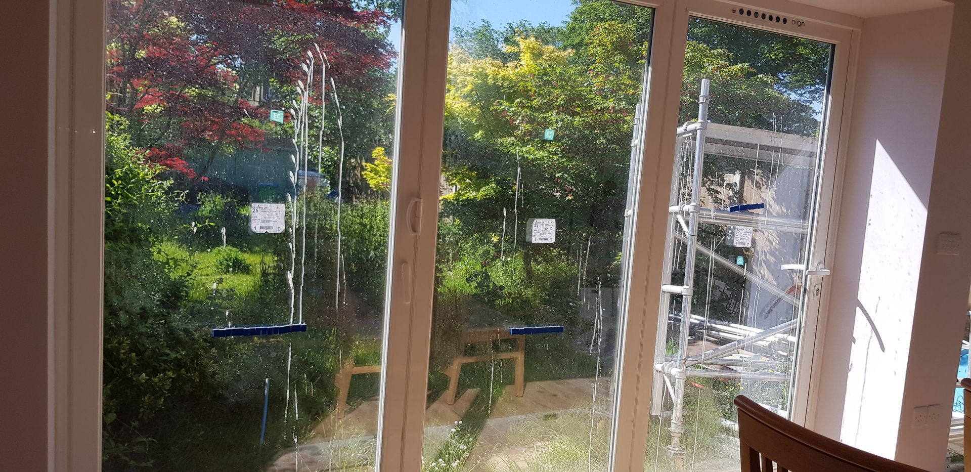 A view through large glass doors, streaked with builders' muck and sticky labels, looking into a garden filled with greenery. A scaffolding structure outside signals ongoing construction, highlighting the need for an After Builders Clean to restore clarity and freshness.