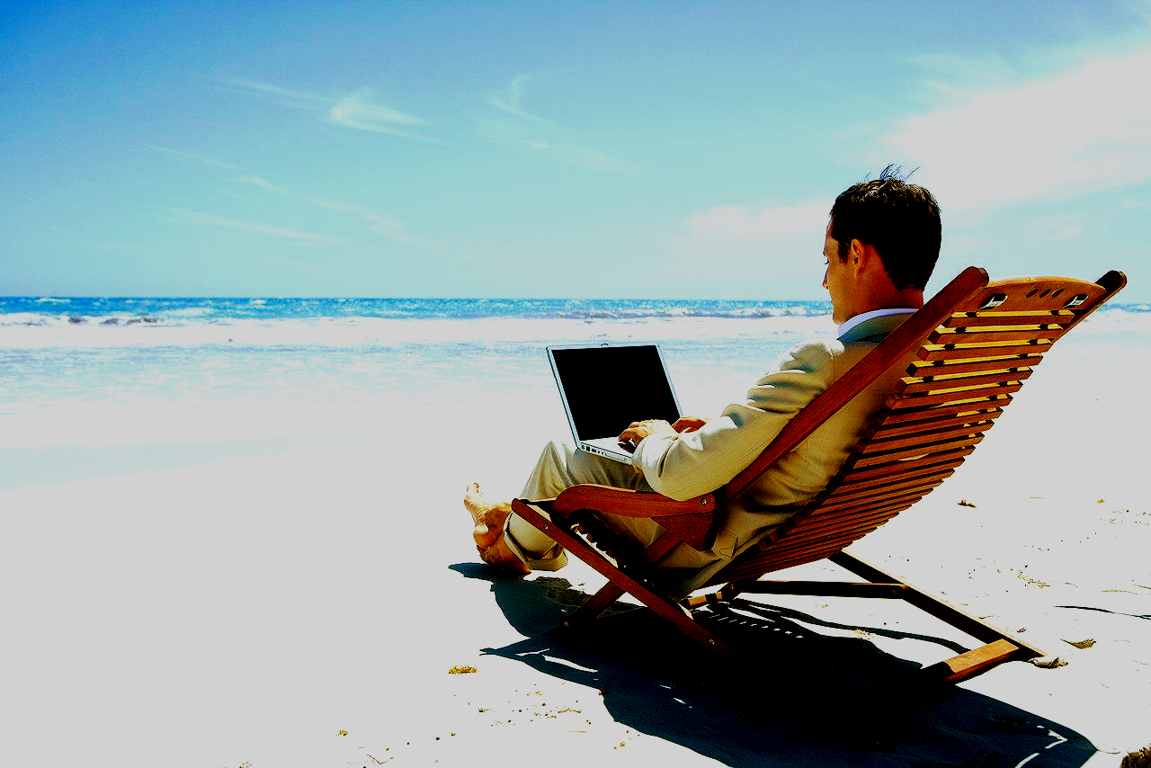 Man in tan suite, no shoes, sitting on a wooden lounge chair on the beach, and working on his laptop.