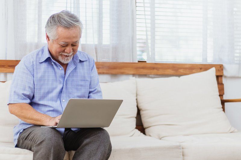Older Asian man in blue shirt, studying on a couch with his laptop in his lap.