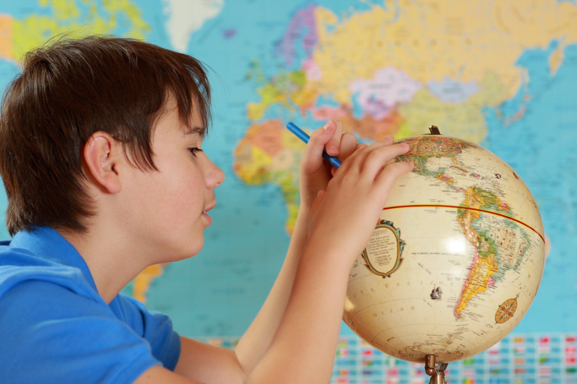 Young boy in blue shirt, with blue pen in hand, trying to locate countries on a globe